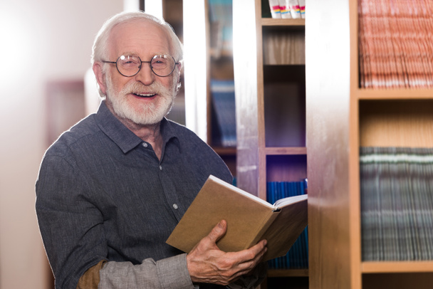 smiling grey hair librarian holding book and looking at camera - Фото, изображение