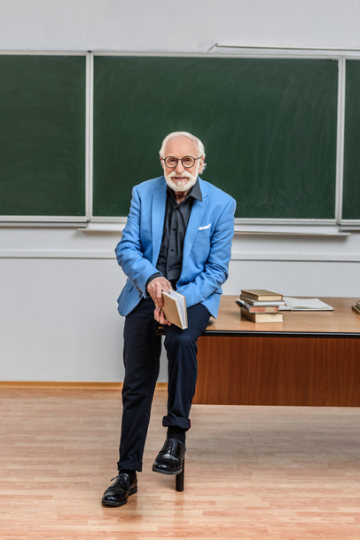 grey hair professor sitting on table in lecture room and holding book - Foto, immagini