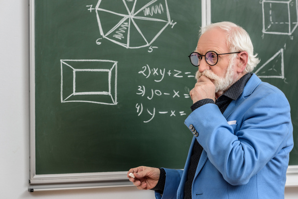 pensive grey hair professor standing at blackboard with piece of chalk - Фото, изображение