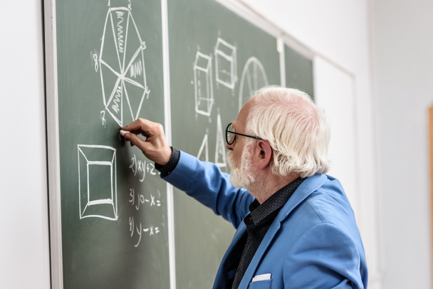 Zijaanzicht van grijze haren professor tekenen met stuk krijt op blackboard - Foto, afbeelding