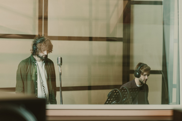 band of two musicians recoding song behind glass at studio - Photo, Image
