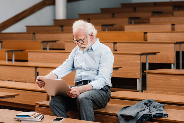 professore di capelli grigi seduto sulla scrivania in aula vuota e utilizzando il computer portatile
 - Foto, immagini