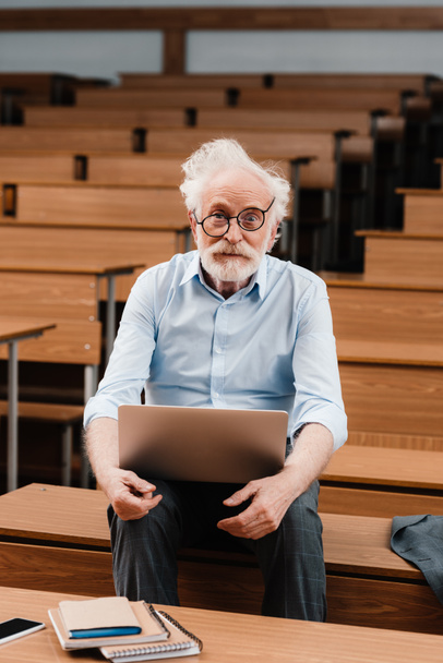 senior lecturer in empty lecture room holding laptop and looking at camera - Fotoğraf, Görsel