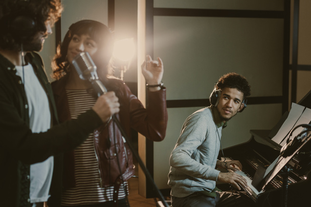 νεαρό ζευγάρι εκτελεί τραγούδι με μικρόφωνο ενώ άτομο παίζει πιάνο πίσω από - Φωτογραφία, εικόνα