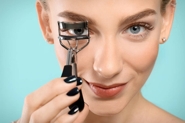 Beautiful young woman with eyelash curler on color background, closeup - Фото, изображение