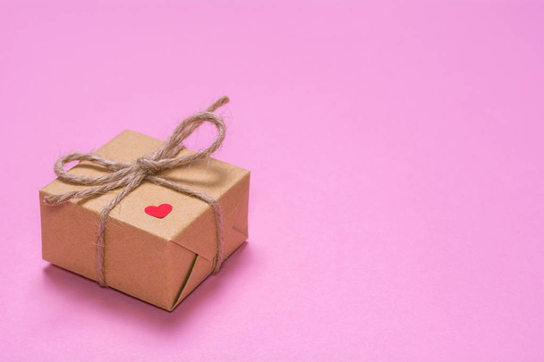 Un regalo envuelto en papel kraft sobre un fondo rosa. Corazón rojo de papel en una caja de regalo
. - Foto, imagen