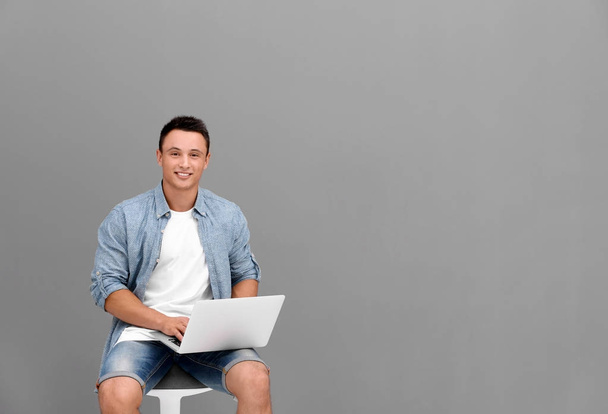 Young man with laptop sitting on chair near grey wall - Фото, зображення