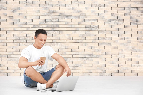 Young man with laptop and cup of coffee sitting on floor near brick wall - Foto, immagini