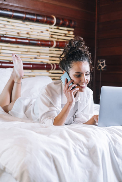 Pretty woman with phone and laptop in bed - Photo, Image