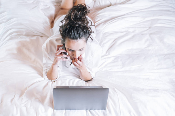 Mooie vrouw met telefoon en laptop in bed - Foto, afbeelding