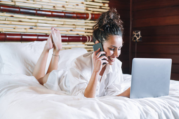 hübsche Frau mit Telefon und Laptop im Bett - Foto, Bild