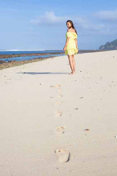 Vertical portrait of beautiful smiling girl in yellow dress walking away along the beach and her footprints - Fotó, kép