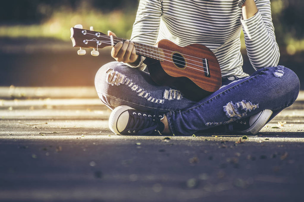 Asian teen girl wearing jeans and T-shirt playing ukulele is within the park. She was sitting on the corridor in the warm sunlight shines from the back with copy space. - Фото, изображение