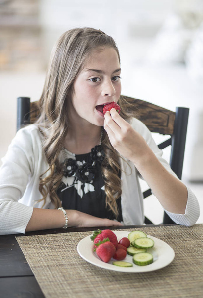 Cute girl sitting at the dinner table eating a healthy plate of fruits and vegetables. She is developing good eating habits eating strawberries and cucumber - Photo, image