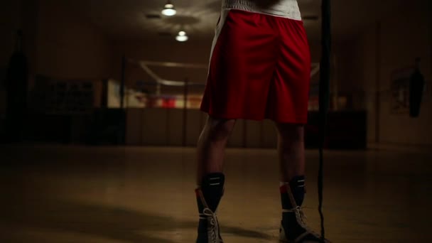 Ein aufgepumpter Mann in Boxstiefeln und kurzen Hosen in der Turnhalle vor dem Hintergrund eines Boxrings trägt Bandagen an den Händen. - Filmmaterial, Video