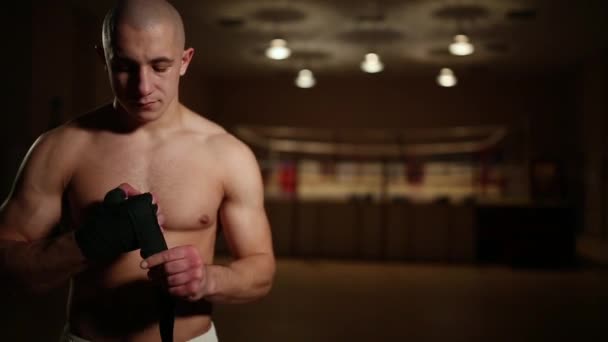 Um homem bombeado de botas de boxe e shorts no ginásio contra o fundo de um anel de boxe veste bandagens em suas mãos
. - Filmagem, Vídeo