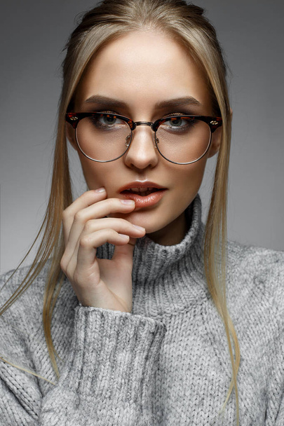 A young blonde girl with straight hair and gentle makeup. Beautiful model in a winter sweater and with glasses. Shining skin. Beauty of the face. Photo is taken in the studio. Cozy image - Photo, Image