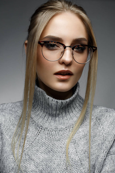 A young blonde girl with straight hair and gentle makeup. Beautiful model in a winter sweater and with glasses. Shining skin. Beauty of the face. Photo is taken in the studio. Cozy image - 写真・画像