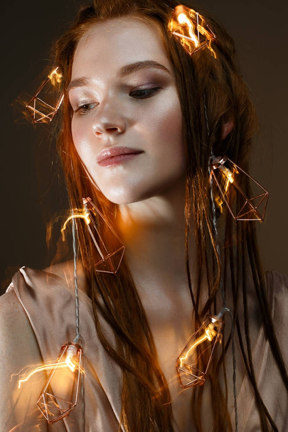 A young girl with red hair and with a luminous garland. Beautiful model with shining bulbs and lights. Gentle makeup. Festive new year's image. Beauty of the face. Photo is taken in the studio. - Photo, Image