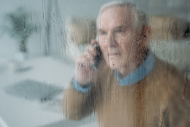 За дождливым стеклянным видом пожилого человека, делающего телефонный звонок
  - Фото, изображение