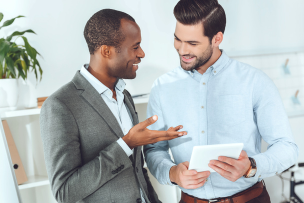 smiling african american and caucasian businessmen talking on office and using tablet - Photo, Image