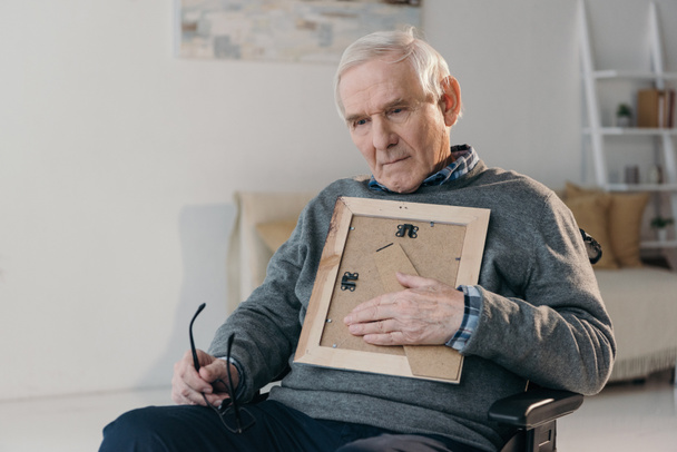 Hombre reflexivo mayor sentado en la silla y sosteniendo el marco de fotos viejo
 - Foto, imagen
