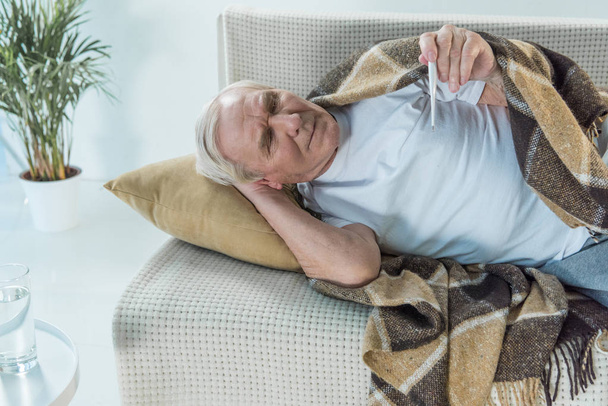 Senior sick man covered in plaid lies on sofa and checks thermometer - Photo, Image