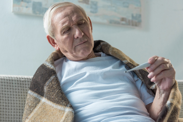 Uomo anziano malato coperto di plaid seduto sul divano e controllare il termometro
 - Foto, immagini
