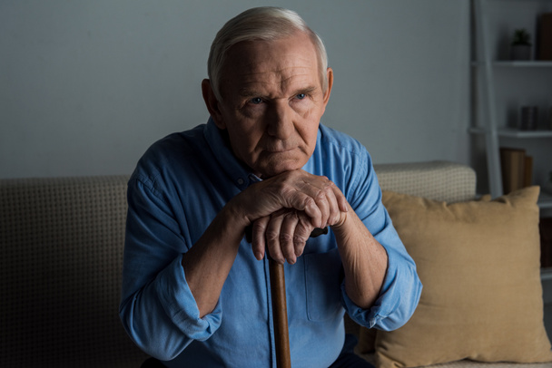 Tired senior man leans on a cane while sitting on sofa - Photo, Image