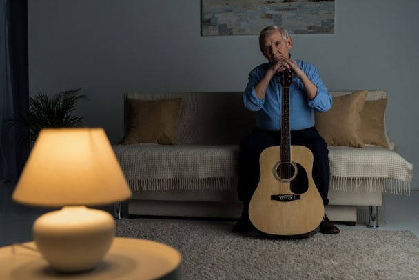 Vanhempi itsevarma mies pitää akustista kitaraa istuessaan sohvalla huoneessa
 - Valokuva, kuva