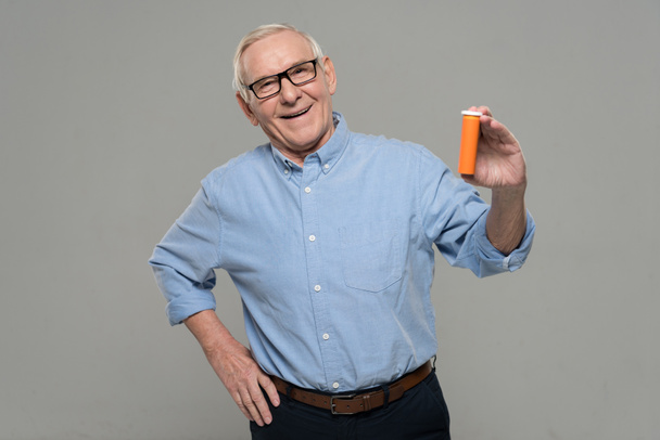 Confident senior man holds blank bottle of pills isolated on gray background - Photo, Image