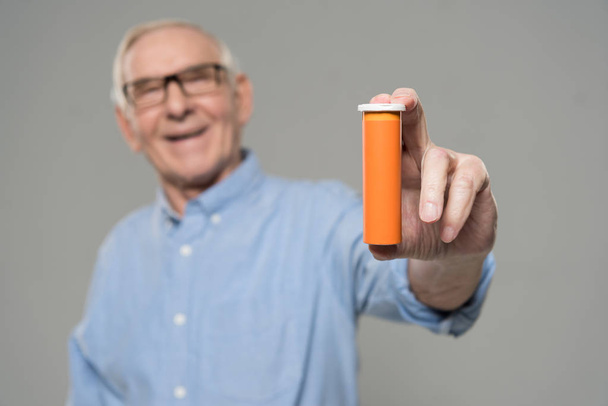 Starší usměvavý muž drží prázdná láhev pilulek izolované na šedém pozadí - Fotografie, Obrázek