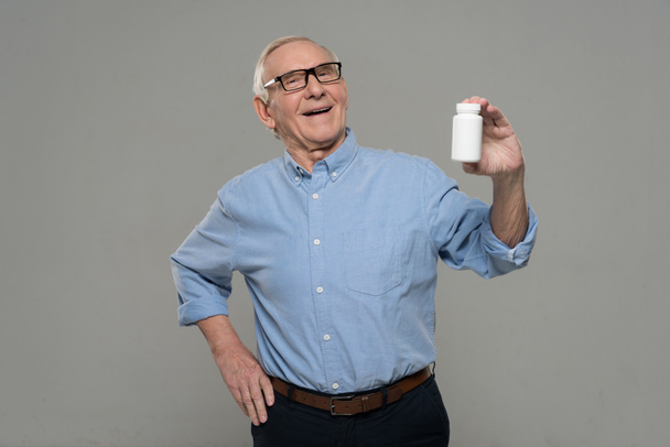 Старший улыбающийся мужчина держит белую бутылку таблеток изолированы на сером фоне
 - Фото, изображение
