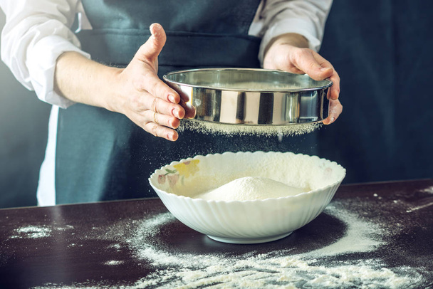 The chef in black apron sifts the flour through a sieve to prepare the dough for pizza - Foto, Imagem