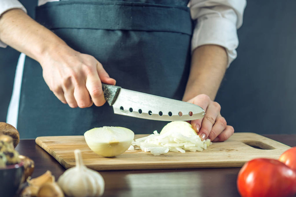 Шеф-повар в черном фартуке режет лук ножом. Концепция экологически чистых продуктов для приготовления пищи
 - Фото, изображение