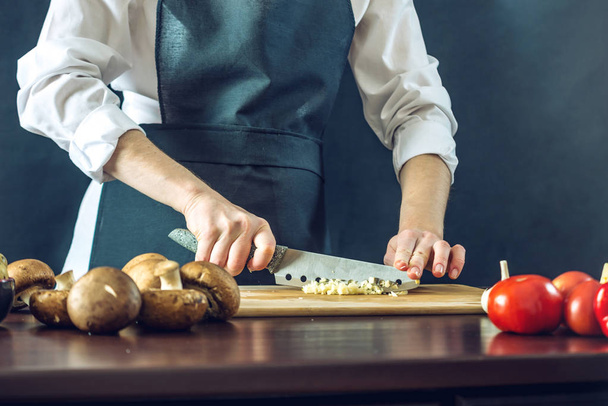 O chef de avental preto corta o alho com uma faca. Conceito de produtos ecológicos para cozinhar
 - Foto, Imagem