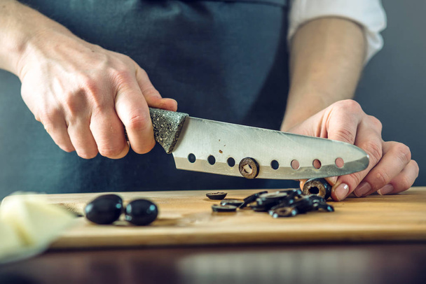 The chef in black apron cuts black olives with a knife. Concept of eco-friendly products for cooking - Foto, afbeelding