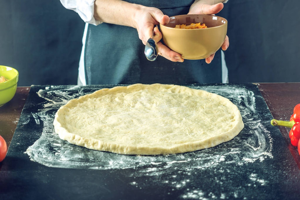 El chef de delantal negro hace pizza con las manos poniendo los ingredientes de la pizza en la mesa
. - Foto, Imagen