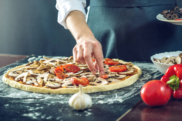 Ο σεφ στην μαύρη ποδιά κάνει πίτσα με τα χέρια του να βάζει τα υλικά για την πίτσα στο τραπέζι. - Φωτογραφία, εικόνα