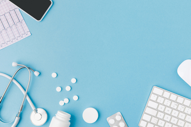 container, verspreide pillen, stethoscoop, cardiogram, toetsenbord en smartphone geïsoleerd op blauwe achtergrond    - Foto, afbeelding