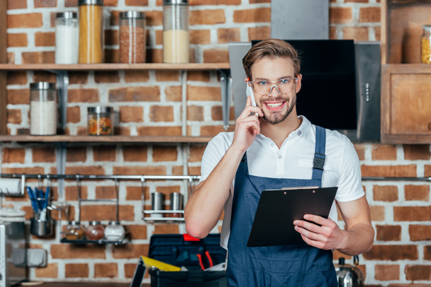 young repairman with clipboard talking by smartphone and smiling at camera in kitchen  - Photo, Image