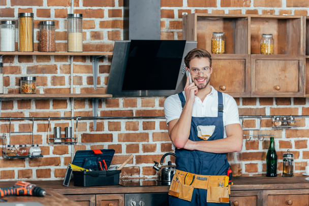 handsome young repairman talking by smartphone and smiling at camera in kitchen  - Photo, Image