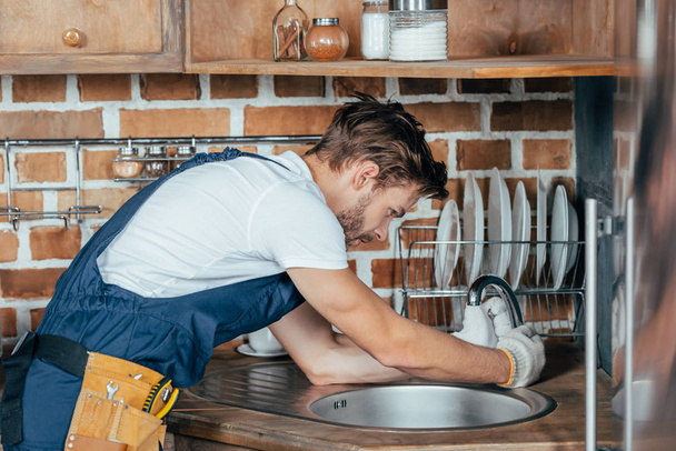 Вид сбоку молодого разнорабочего, ремонтирующего раковину на кухне
 - Фото, изображение