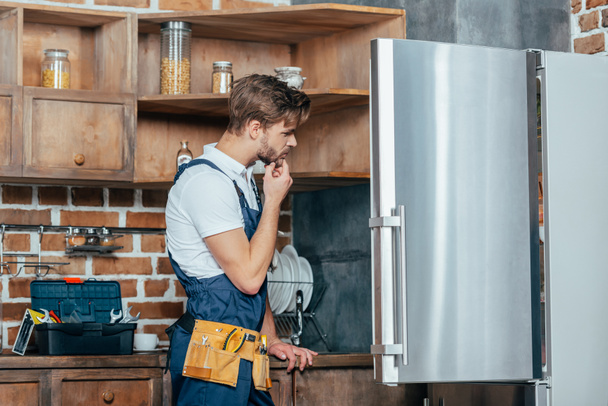 молодой ремонтник с инструментальным поясом смотрит на сломанный холодильник
 - Фото, изображение