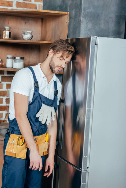 tired young foreman with tool belt leaning at broken refrigerator - Fotografie, Obrázek
