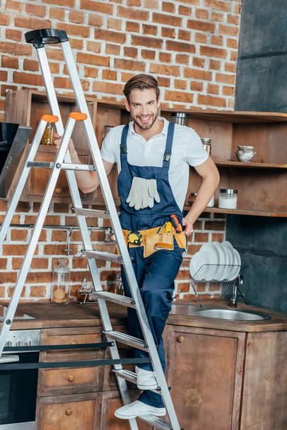 pohledný mladý opravář stojí na žebříku a usmívá se na kameru v kuchyni - Fotografie, Obrázek