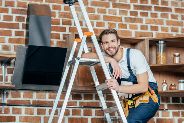 handsome young foreman standing on ladder and smiling at camera while fixing kitchen hood  - Photo, Image