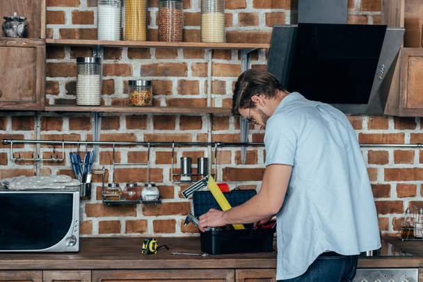 Junger Mann sucht in Werkzeugkiste mit Werkzeug, während er in Küche arbeitet - Foto, Bild