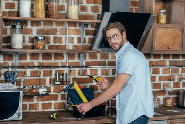 молодий чоловік в окулярах бере інструменти з коробки інструментів і дивиться на камеру на кухні
 - Фото, зображення