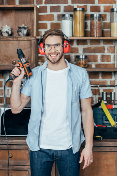 handsome young man in eyeglasses and earmuffs holding electric drill and smiling at camera - Foto, imagen